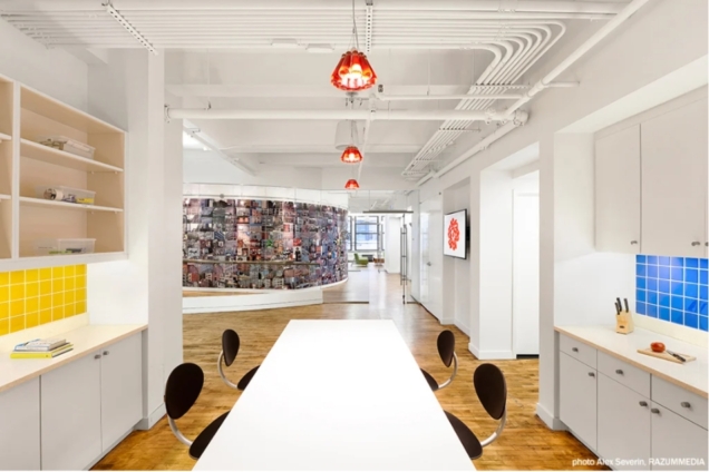 an image of a large space with natural lighting. In the foreground the walls are lined with cabinets and shelves. There is a long table with chairs. In the background glass doors lead to an entry way with a curved wall and the entrance to another room.