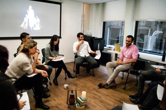 image of eight individuals sitting in chairs in a circle. the individuals hold papers. Some are writing and listening. IN the background, a presentation is projected onto a screen.