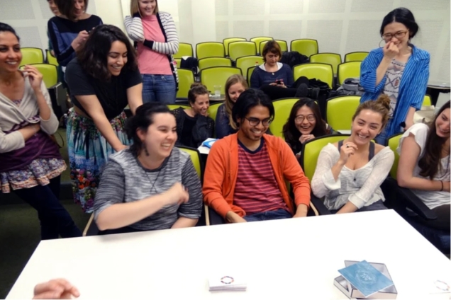 image shows a group of fourteen smiling individuals seated in lime green auditorium chairs. The group of people are looking at a table in front of them that shows various prototypes including a deck of cards.