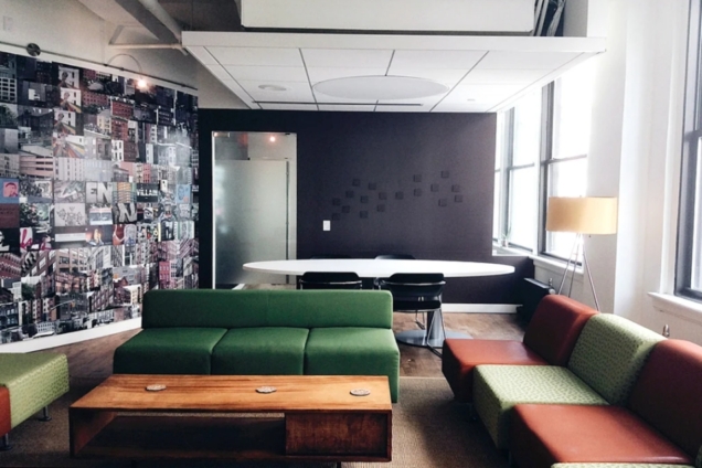 image of lounge space showing walls of images of NYC city-scape and a chalkboard wall. In the background is a conference table and chairs. In the foreground there are green and orange couches around a wooden coffee table