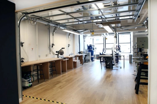image shows large open floor plan work space with wooden flors, wooden tables, and stools tucked underneath. There are various pieces of equipment