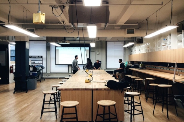 image of large workspace with wooden tables and stools. there are four individuals moving in the background,, working in the space. The wall is lined with a screen projector and wooden shelving.