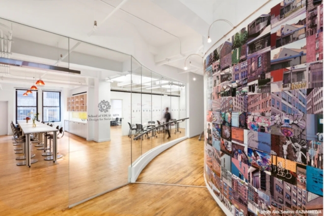Image of entrance to DSI. Shows a well-lit space with hardwood floors. One door is propped open leading to a long, high table and chairs. In the background there are more tables and chairs and a person walking by. There is another curved ramp entrance along a wall with a patchwork of images of NYC.