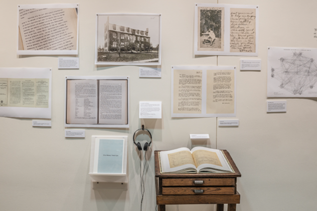 An image of a book on a wooden pedestal in front of a white wall with various posters on it. Each of the posters has historical images or documents depicted on them.