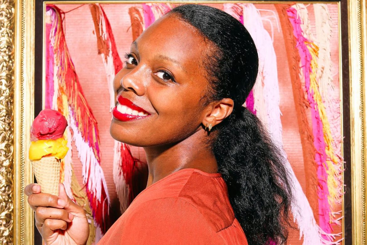 A close-up photo of Lokelani Alabanza in a salmon-colored top. She is looking over her left shoulder and smiling at the camera while holding an ice cream cone. The cone has two scoops on it: one yellow and one red. The background is various shades of pink.