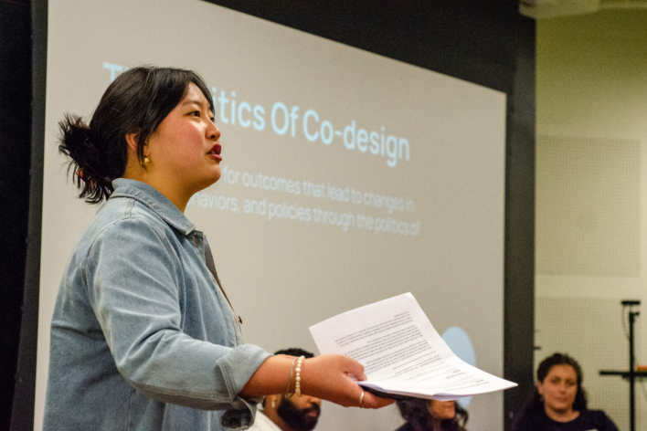 image of individual speaking while holding notes; in the background is a screen and seated individuals