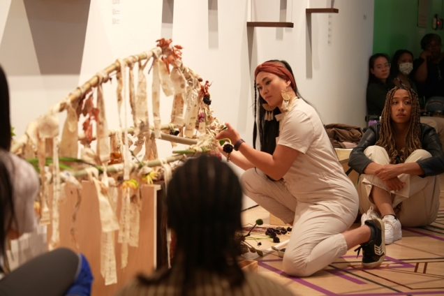 An image of Kimberly Tate in a white jumpsuit. They are arranging an ornate artifact with hanging tassels and paper talismans.
