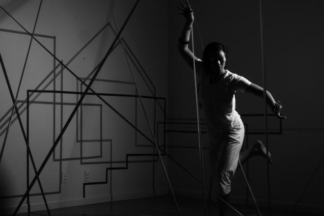 A black and white image of an Asian woman dancing while shrouded partially in shadows. Behind her is a complex arrangement of lines in intersecting patterns.