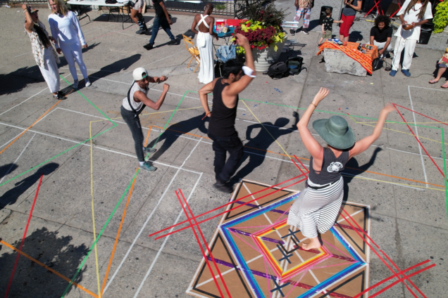 People dancing over a complex arrangement of multicolored lines drawn on pavement. There are people near them playing instruments, and other people standing around having conversation in the background.
