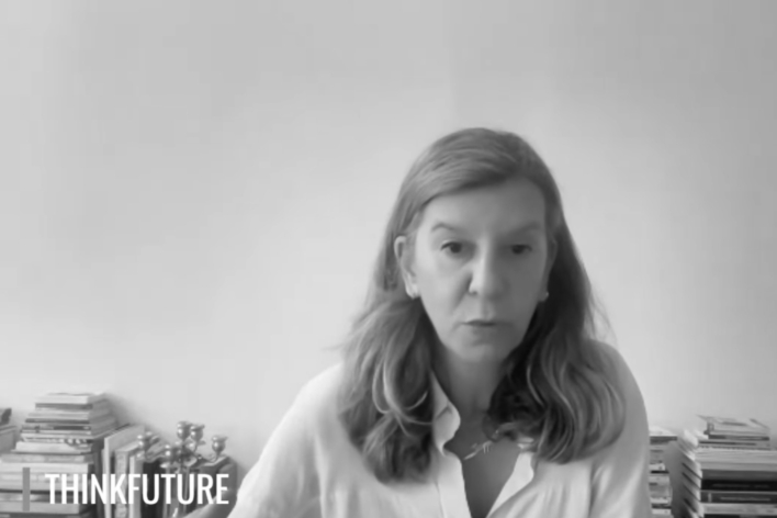 A black and white screenshot of a woman speaking to the camera. In the bottom left corner there is a logo in white text that reads: