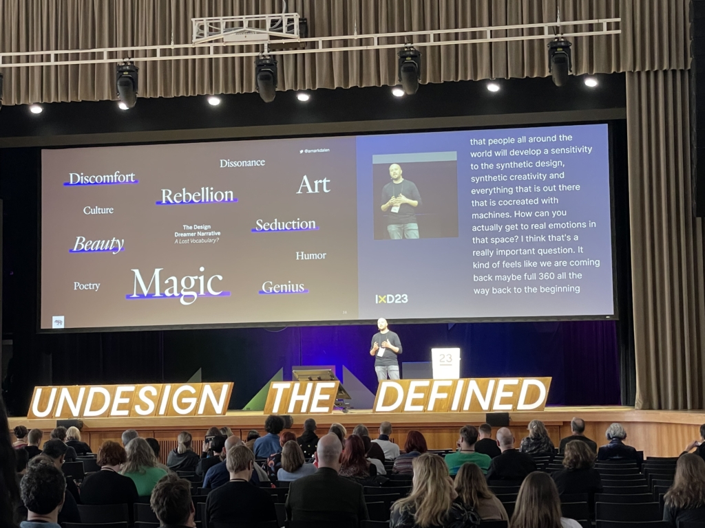 A man in a black shirt and jeans giving a presentation on a stage. Behind him are various keywords in white text on a black background. He is standing behind a long sign that reads "Undesign the Defined"