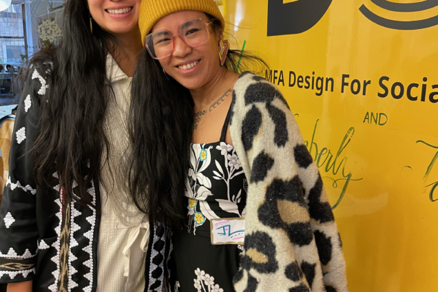 Two women standing side by side smiling at the camera in front of a yellow wall. The woman on the left is wearing all white with a long, black cardigan. The woman on the right is wearing a yellow hat, a black and white patterned dress, and a cow print patterned sweater.