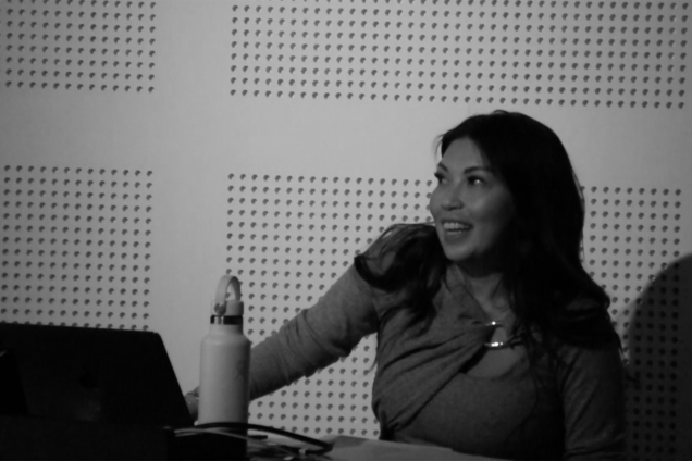 A black and white image of an Asian woman standing at a podium giving a presentation. She is smiling and looking to the left of the image.
