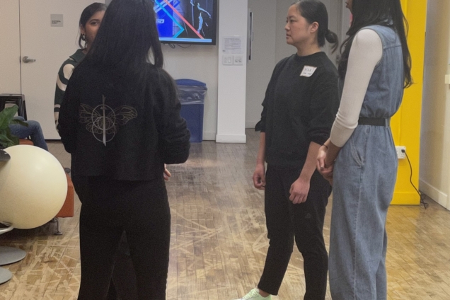 Four women standing in a circle conversing with each other. 3 of the women are wearing comfortable, black sportswear. One of the women is wearing a denim jumpsuit with a white long-sleeved shirt underneath it.