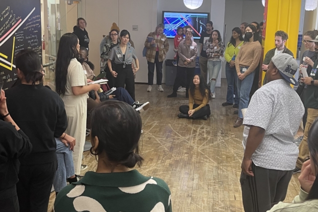 A room of people congregated in a circle around a speaker. The speaker is a woman wearing a white jumpsuit.
