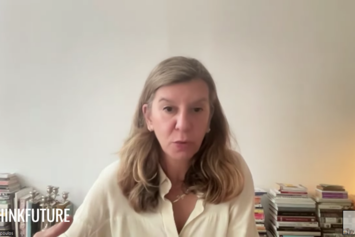A screenshot of a blonde woman speaking to the camera. In the bottom left corner there is a logo in white text that reads:
