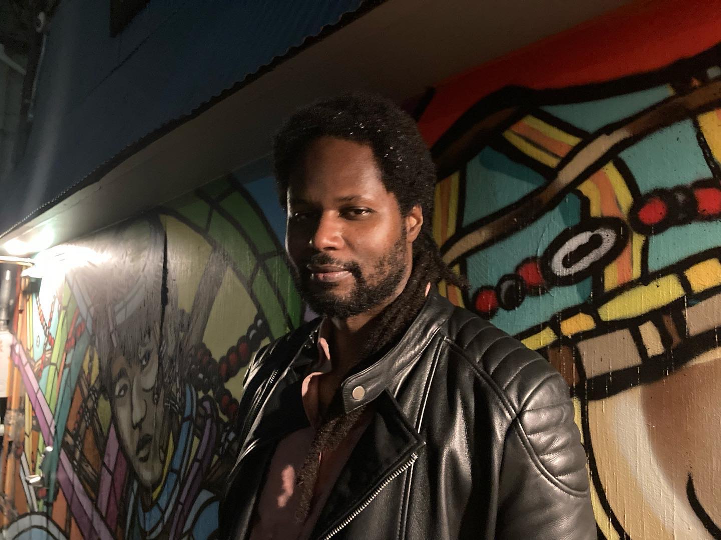 A photograph of a Black man wearing a leather jacket smiling at the camera. He is standing in front of a wall of colorful art.