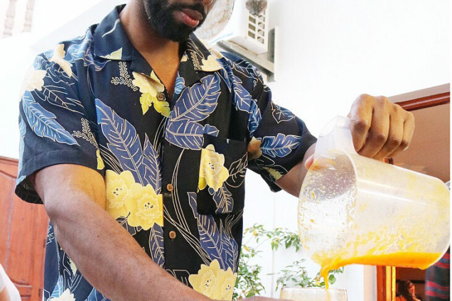An image of a man in a multicolored navy and blue top wearing a hat. He is pouring a cup of juice from a blender into a glass on a wooden cutting board.