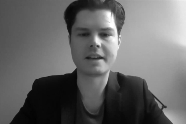 A black and white screenshot of a man wearing a black blazer looking at the camera.