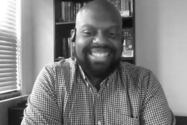 A black and white screenshot of a man wearing a button down shirt looking at the camera.