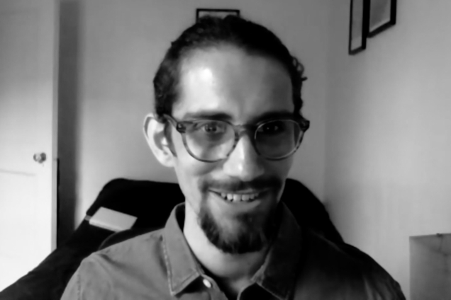A black and white screenshot of a man wearing a grey shirt and glasses looking at the camera.