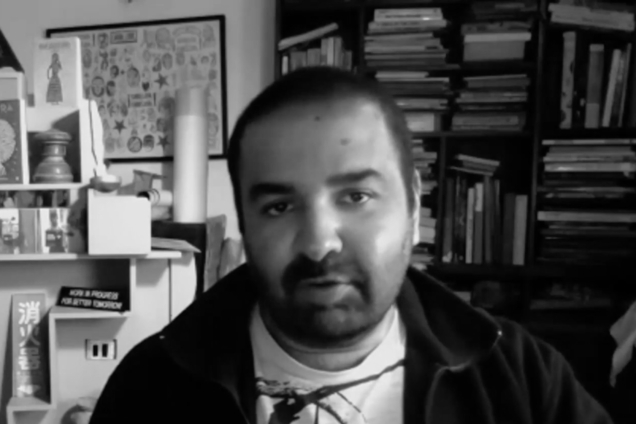 A black and white screenshot of a man wearing a sweater and a t-shirt looking at the camera.