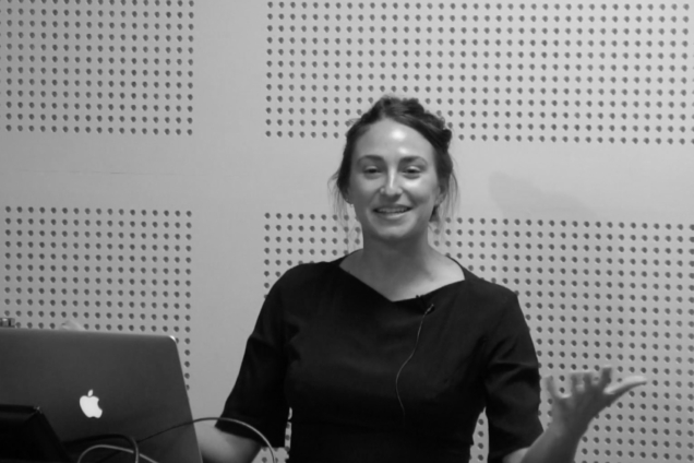 A woman standing at a podium wearing a black top, gesturing to the audience.