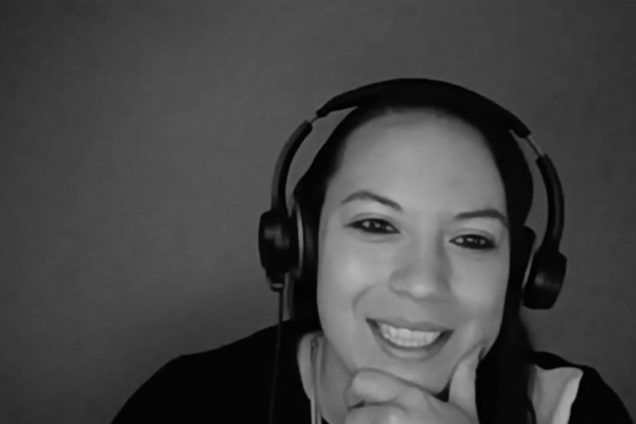 A black and white screenshot of a woman with headphones and a black t-shirt looking at the camera.