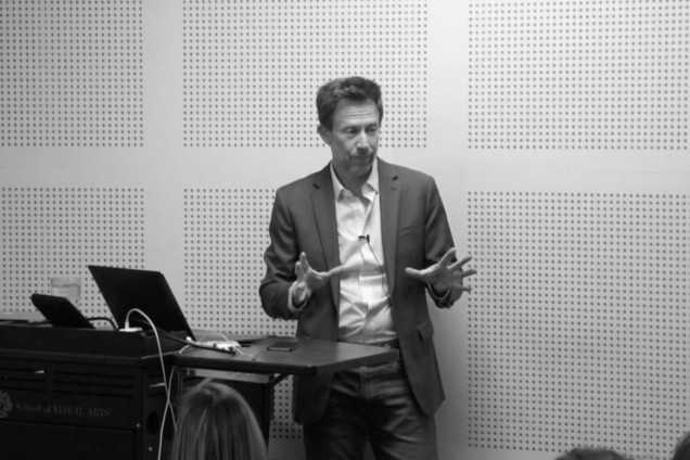 A man in a suit, giving a presentation at a podium.