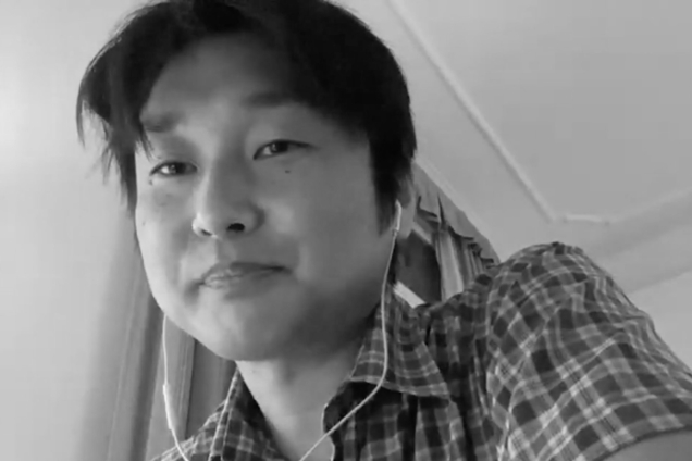 A black and white screenshot of a man wearing Apple earbuds and a button down shirt looking at the camera.