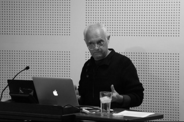 A black and white image of a man in a black button down shirt giving a presentation.