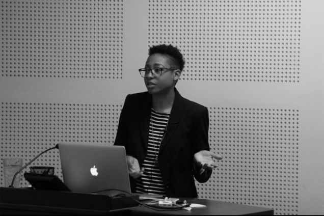 A black and white image of a woman in a black jacket standing at a podium and giving a presentation.