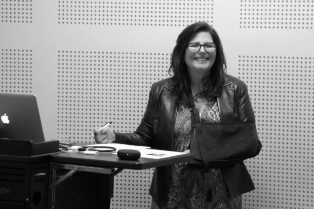 A woman in a leather jacket wearing an arm sling, smiling to an audience.