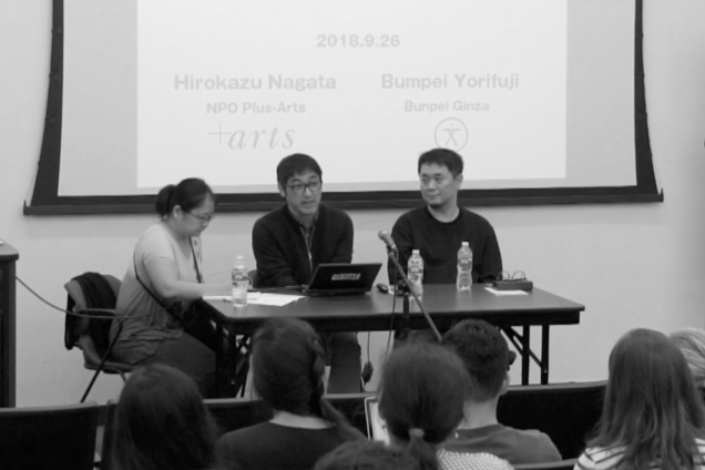 Two men and a woman sitting at a desk in front of a screen that has a presentation on it.