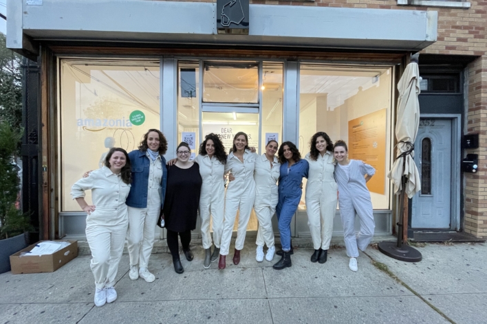 An image of a group of women standing in front of a store front posing for a photo together. All of the women are wearing painter's jumpsuits, except for one who is wearing a black dress and another wearing a denim jumpsuit.