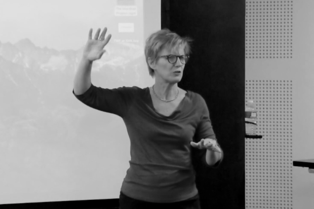 A woman in a dark long-sleeved top speaking to an audience.