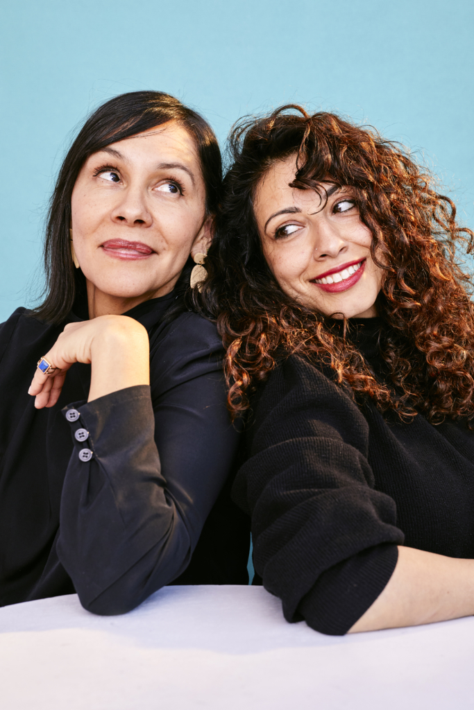 An image of two women wearing black posing over a blue background.