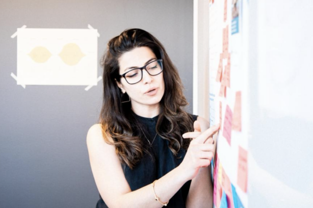 Woman with dark hair, olive skin and glasses stands in front of a whiteboard with post-its