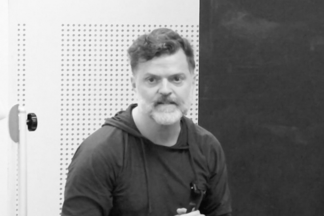 A black and white image of a man sitting, giving a presentation on the podium.