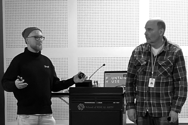 A young man in a sweater presenting. Another man looking at the presenter.