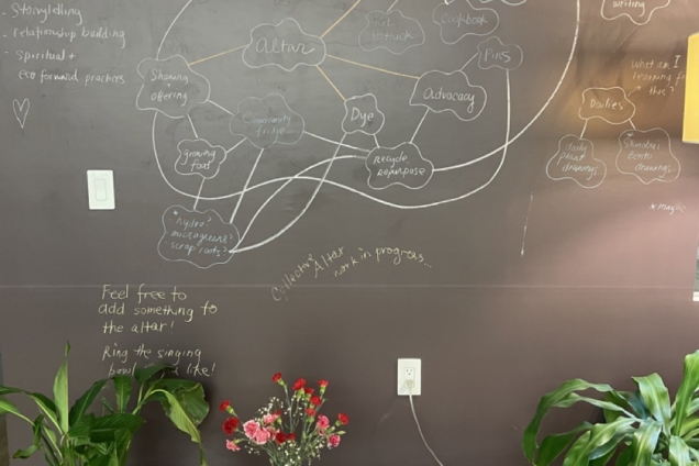 Show Love Thru Food wooden altar in front a chalkboard with two green plants on either side.