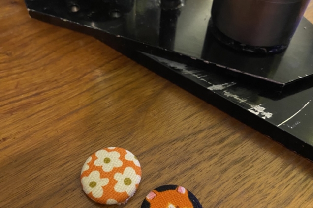Two pins on a wooden table, one with white flowers on a orange background and one with an illustration of an orange dog.