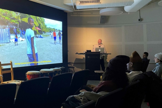 The documentary film Junior on the screen with drummer Markus Schwartz at the podium in a darkened auditorium.