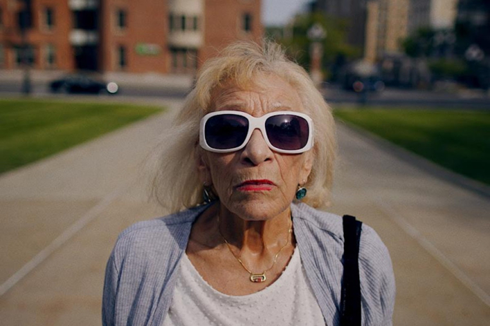 An image of Hilda O. with blond hair, red lipstick, and white sunglasses, staring at the camera not smiling.