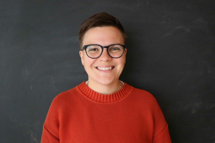 A person with brown short hair and glasses in an orange sweater.