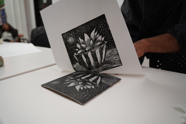 A hand peeling back a paper with a black and while linocut of a plant.