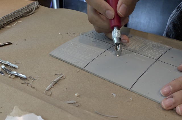 Grey linoleum squares with a hand holding a linocut tool.
