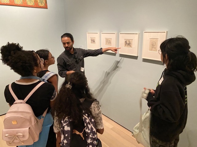 A man with dark hair is pointing to a set of 4 framed images on a blue wall to a group of teen girls.