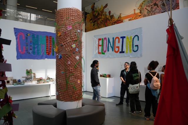 MoMA Art Gallery with people standing in a community exhibition with a quilted multi-colored sign on the wall that says "BELONGING"